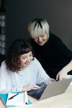 Charming Asian females in home clothes sharing laptop while developing collaborative project together